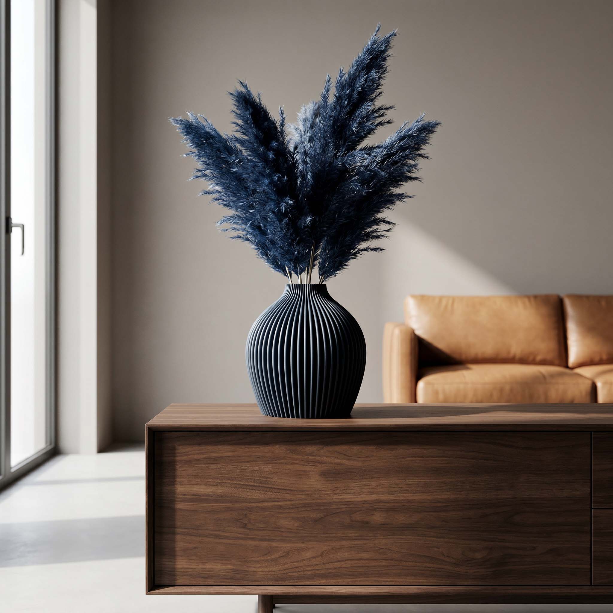 Blauer Federbusch in einer gerippten Vase steht auf einer Holzkommode neben einem Sofa, im Licht am Fenster.